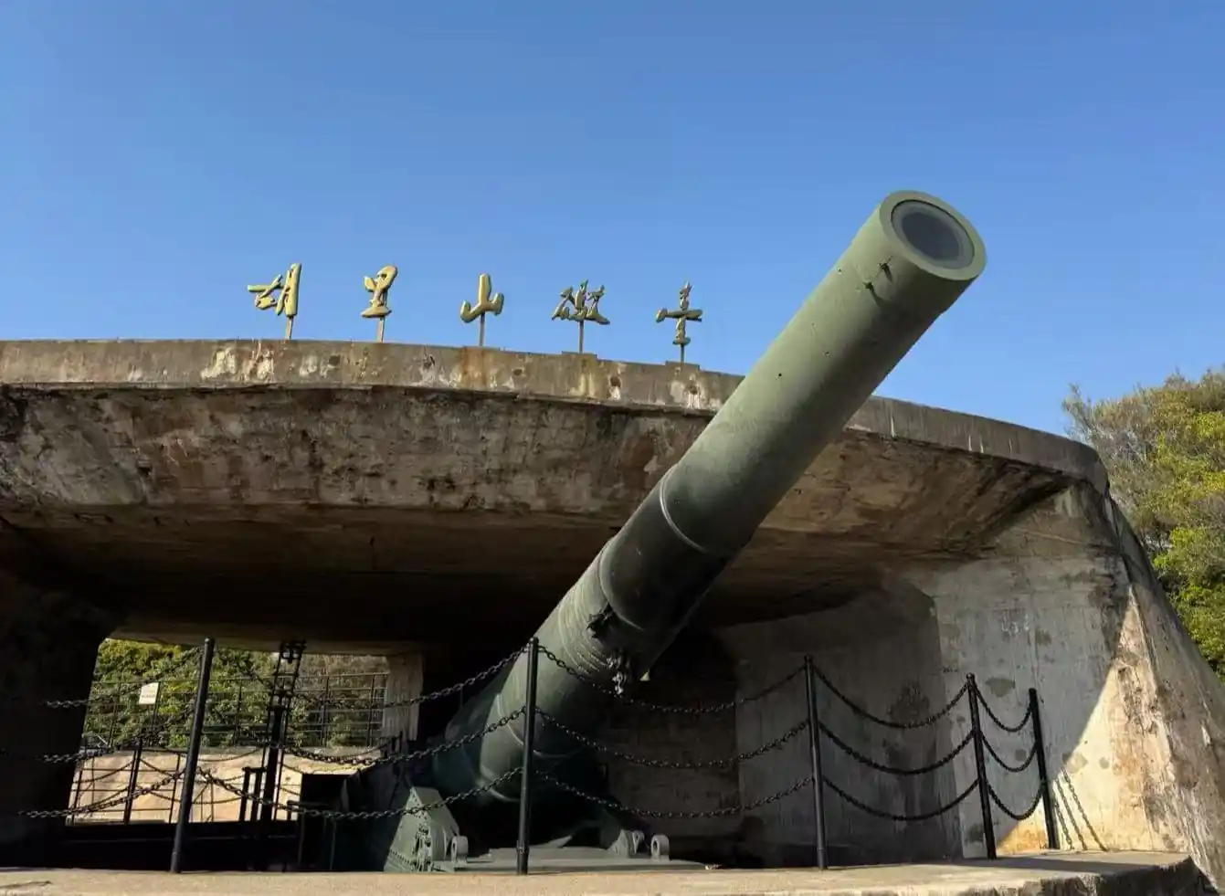 Hulishan Fortress,Xiamen,Chinatour Military cannon protruding from a concrete bunker, angled upward, with blue sky and chain barriers around the display.