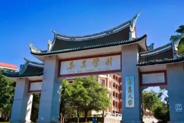 Jimei Educational Village,Xiamen,Chinatour Stone arch gate with traditional Chinese curved roofs, gold sign reading 'The Jimei School Village' and campus buildings visible behind under a clear blue sky.