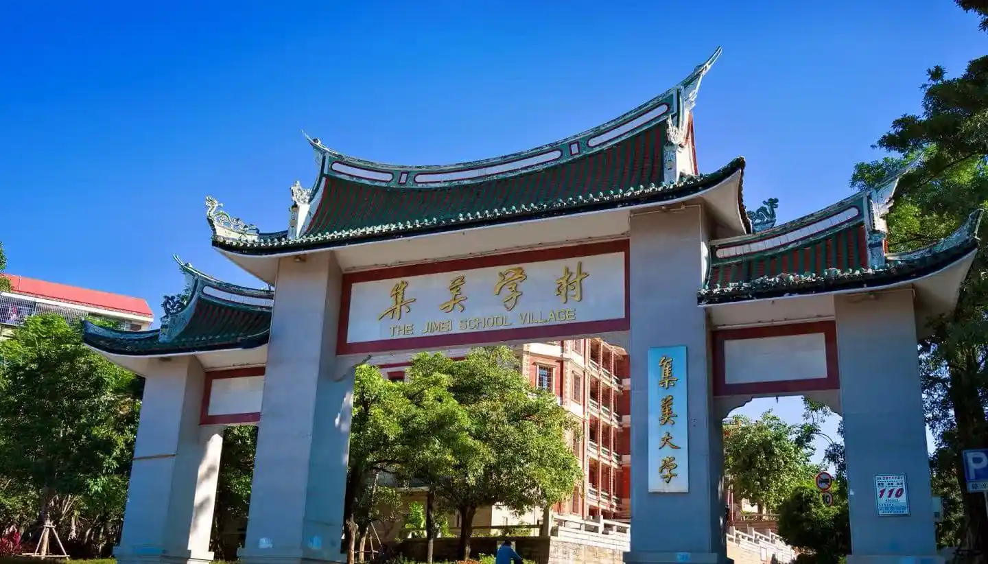 Jimei Educational Village,Xiamen,Chinatour Stone arch gate with traditional Chinese curved roofs, gold sign reading 'The Jimei School Village' and campus buildings visible behind under a clear blue sky.