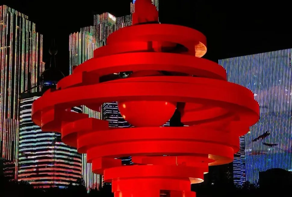 Red spiraling sculpture in a nighttime plaza with tall, brightly lit skyscrapers in the background and city lights.
