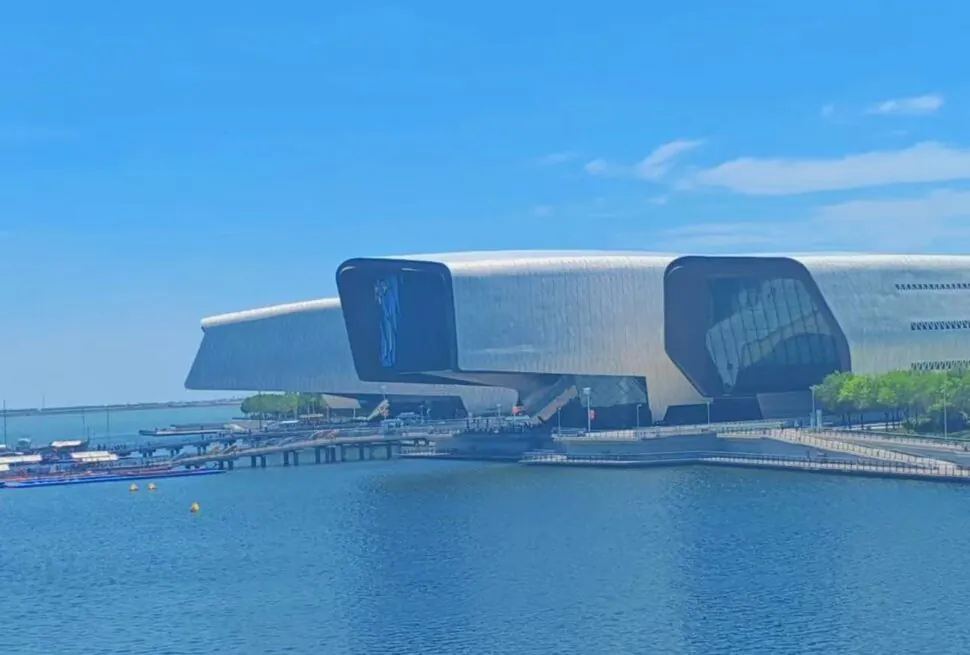 National Maritime Museum,Tianjin,Chinatour Futuristic silver cultural building with curved, wing-like structures along a calm blue waterfront and a wooden boardwalk with boats nearby.