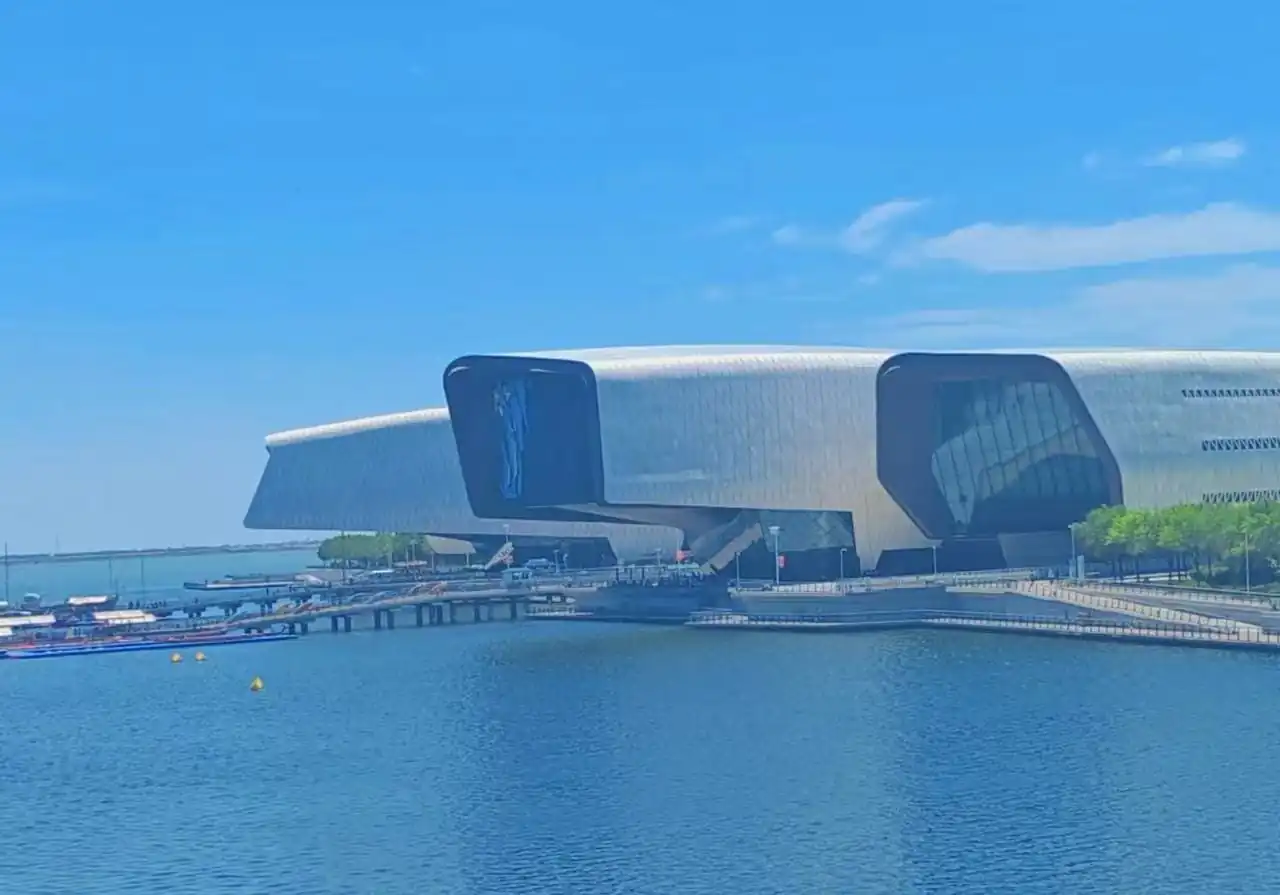 National Maritime Museum,Tianjin,Chinatour Futuristic silver cultural building with curved, wing-like structures along a calm blue waterfront and a wooden boardwalk with boats nearby.
