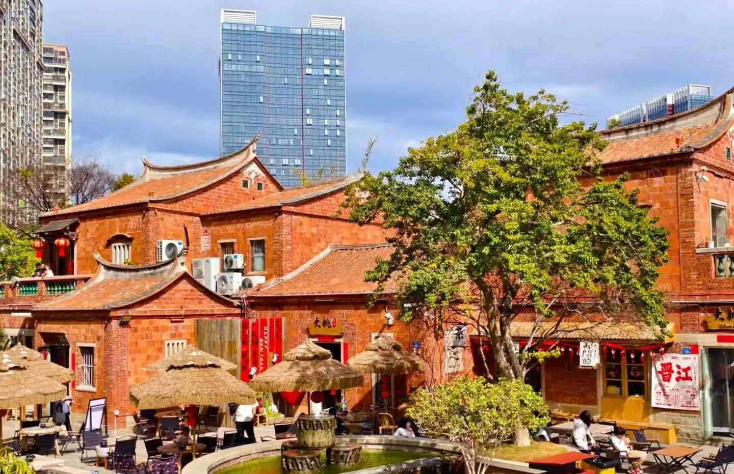 Traditional red-brick Chinese courtyard with thatched umbrellas, outdoor seating, and a stone fountain; modern high-rise in the background with blue sky behind.