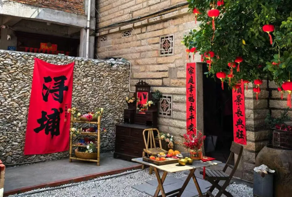Xunpu Village ,Quanzhou,Chinatour Red vertical Chinese New Year couplets hang by a stone wall beside a small wooden altar with flowers and fruit offerings on a folding table outside a doorway.