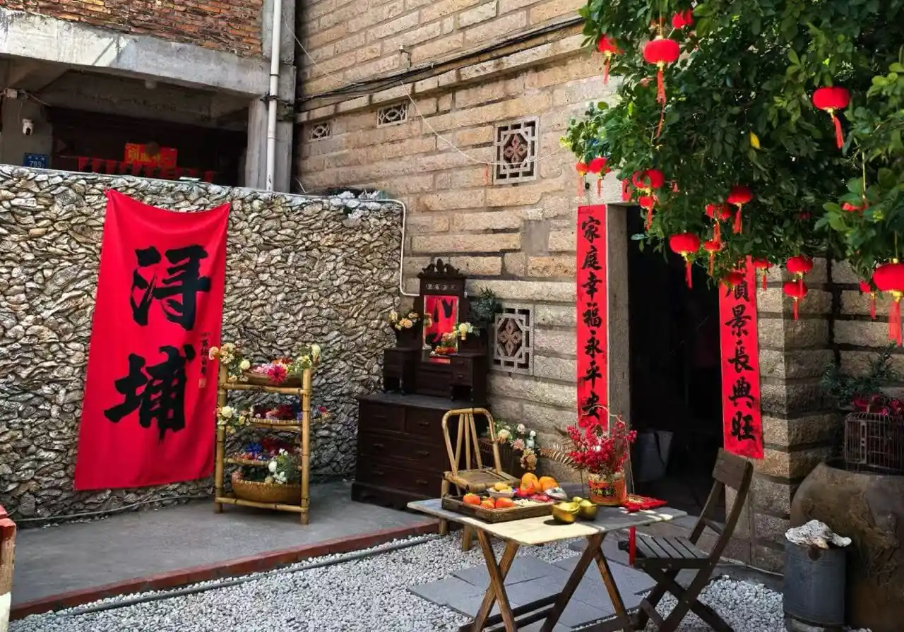 Xunpu Village ,Quanzhou,Chinatour Red vertical Chinese New Year couplets hang by a stone wall beside a small wooden altar with flowers and fruit offerings on a folding table outside a doorway.