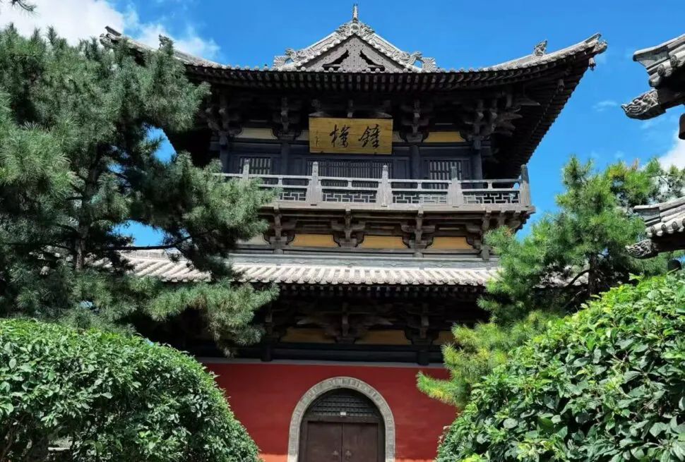 Two-story traditional Chinese temple with ornate curved roofs, a gold signboard, and surrounding pine trees against a blue sky.