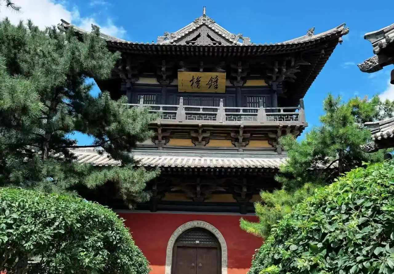 Two-story traditional Chinese temple with ornate curved roofs, a gold signboard, and surrounding pine trees against a blue sky.