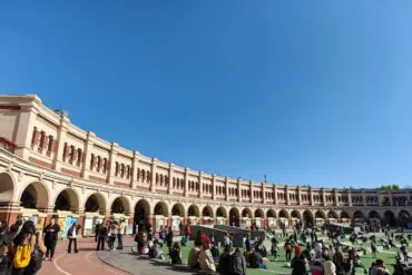 Curved cream-colored arcade with arches surrounds a grassy plaza where people sit and chat on a sunny day.