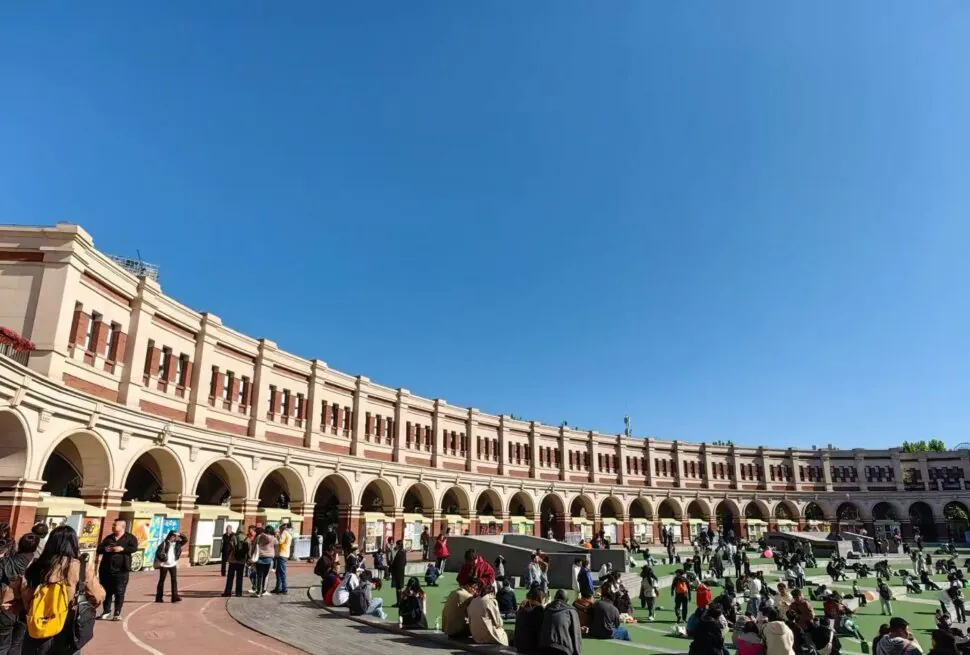 Curved cream-colored arcade with arches surrounds a grassy plaza where people sit and chat on a sunny day.