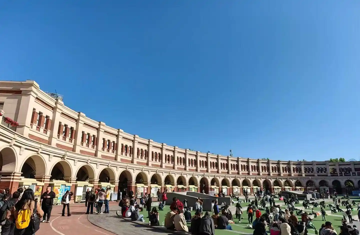 Curved cream-colored arcade with arches surrounds a grassy plaza where people sit and chat on a sunny day.