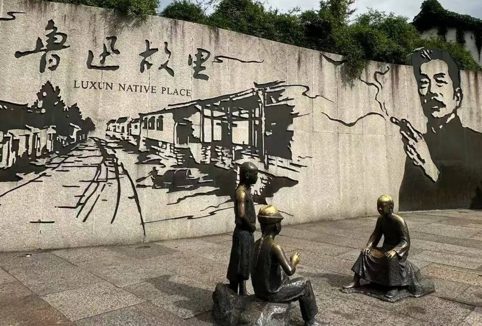 History and Culture of Lu Xun Native Place,Shaoxing,Chinatour Bronze statues of four people in a plaza in front of a concrete wall mural that reads 'Luxun Native Place' with a street scene illustration and a large portrait on the right.
