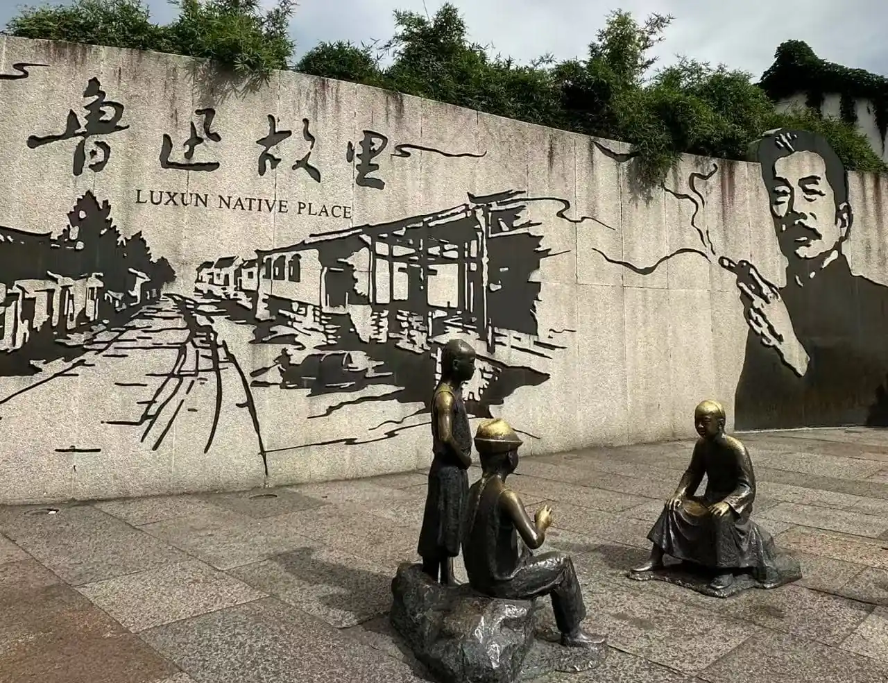 History and Culture of Lu Xun Native Place,Shaoxing,Chinatour Bronze statues of four people in a plaza in front of a concrete wall mural that reads 'Luxun Native Place' with a street scene illustration and a large portrait on the right.