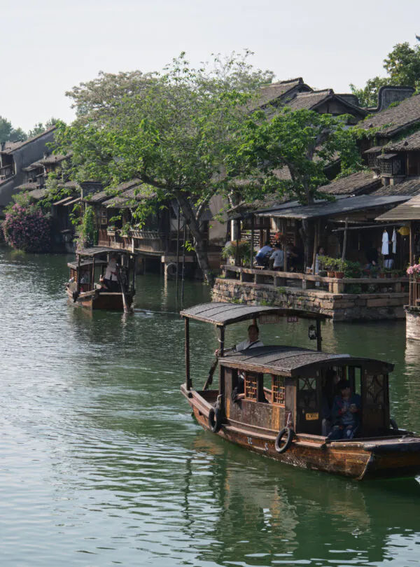 Zhejiang Weather ,Zhejiang,Chinatravel Boats travel along a canal beside traditional wooden houses and a stone bridge with people walking above and along the water's edge, surrounded by trees and plants.