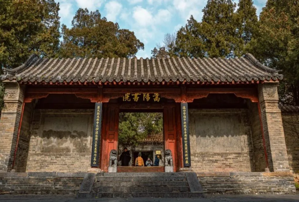 Traditional Chinese temple gate with a tiled roof, red pillars, gold inscription, guardian lions, and visitors standing under the doorway.Songyang Academy,Shaolin & Songshan Area,Henan,Chinatravel