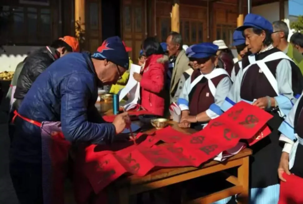 People in blue and white traditional outfits gather around a table as a man signs red banners with Chinese calligraphy outdoors at a festival.Lijiang Old Town,Lijiang,Yunnan,Chinatravel