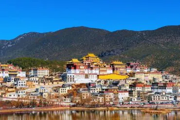 A hillside town with white buildings and yellow-roofed temples under a bright blue sky, beside a calm river.Songzanlin Monastery,Shangri-La,Yunnan,Chinatravel