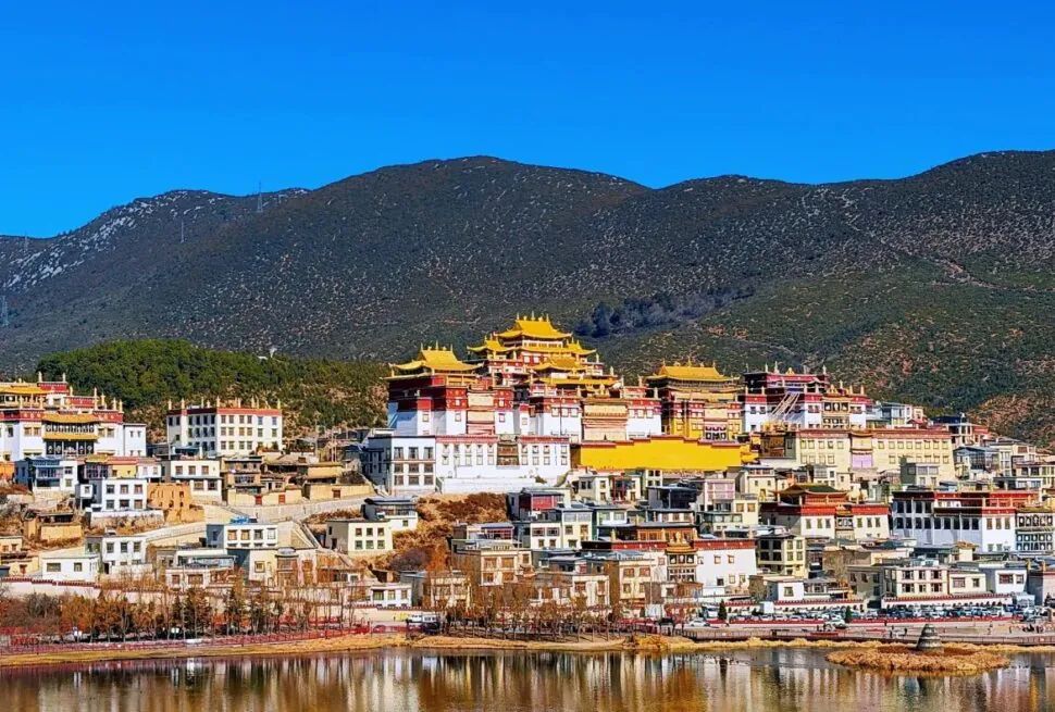A hillside town with white buildings and yellow-roofed temples under a bright blue sky, beside a calm river.Songzanlin Monastery,Shangri-La,Yunnan,Chinatravel