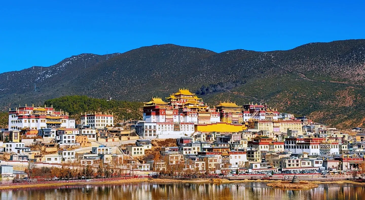 A hillside town with white buildings and yellow-roofed temples under a bright blue sky, beside a calm river.Songzanlin Monastery,Shangri-La,Yunnan,Chinatravel