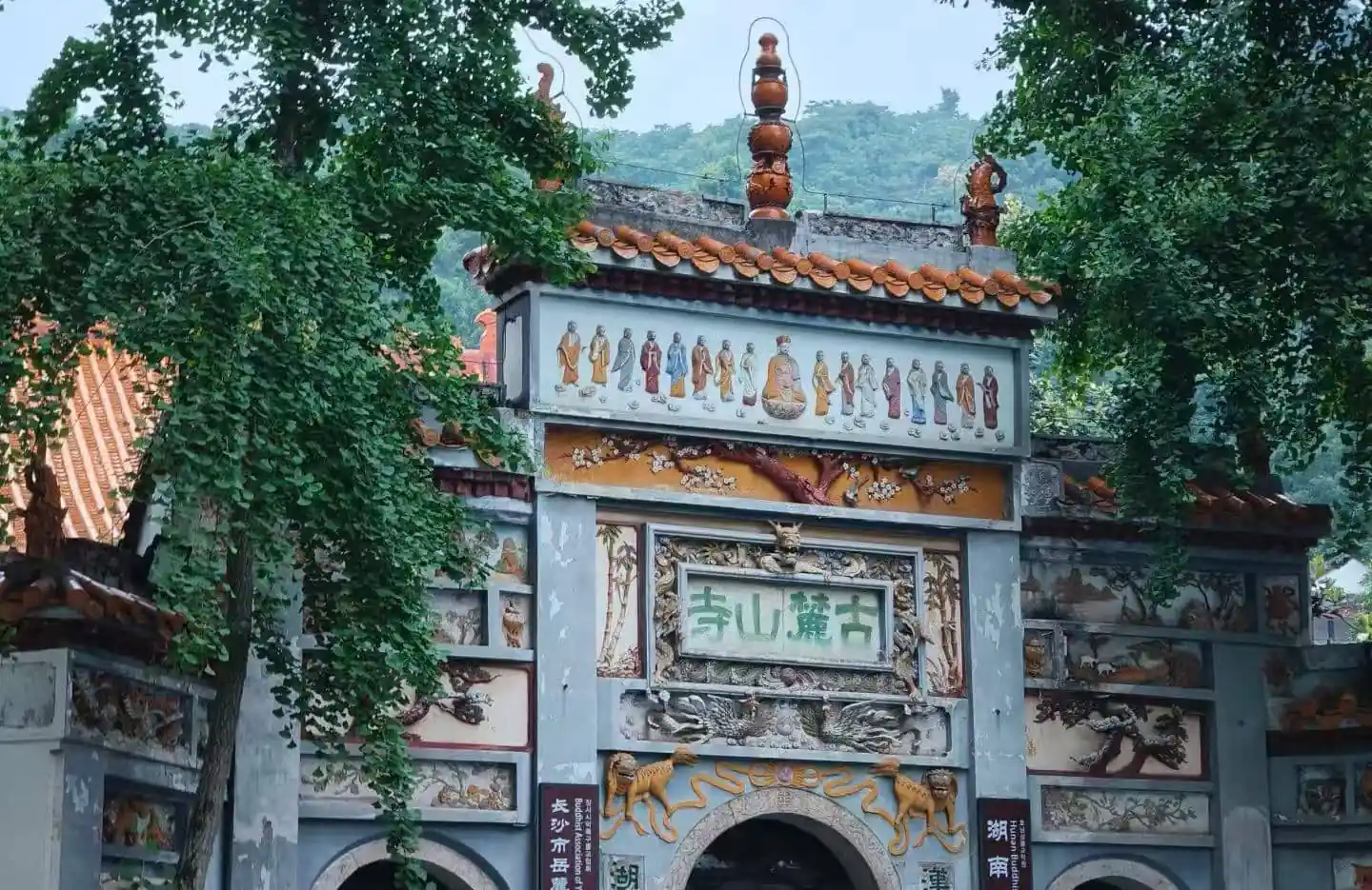 Ornate temple gate with gray stone walls, colorful murals of Buddhist figures, and trees framing the scene.Lushan Templen,Changsha,Hunan ,Chinatravel