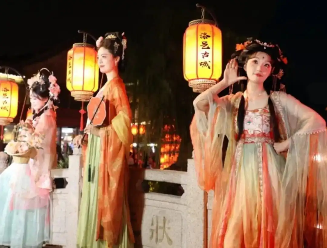 Luoyi Ancient City,Luoyang,Chinatour Three women in colorful traditional Chinese hanfu outfits posing with glowing lanterns at night festival on a stone railing.