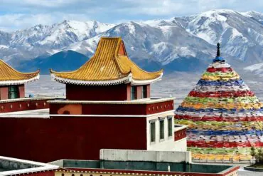 Red and gold-trimmed temple buildings with tiered roofs set against snow-capped mountains and a colorful prayer-stone cone nearby, Tibet.Riyue Mountain Pass,Xining,Qinghai