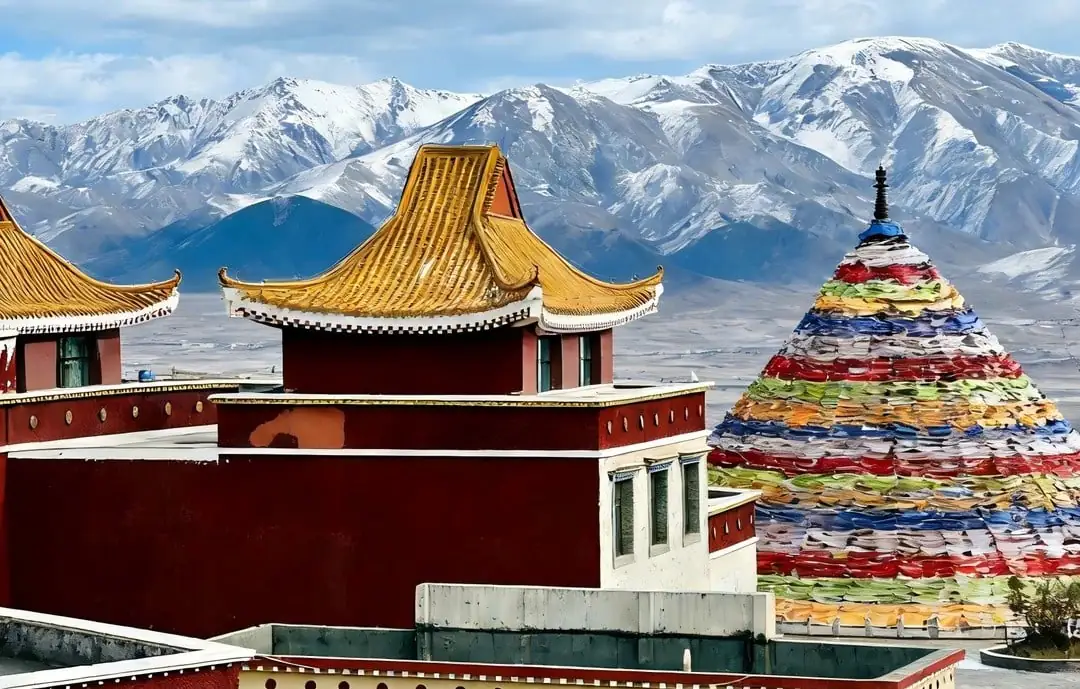 Red and gold-trimmed temple buildings with tiered roofs set against snow-capped mountains and a colorful prayer-stone cone nearby, Tibet.Riyue Mountain Pass,Xining,Qinghai