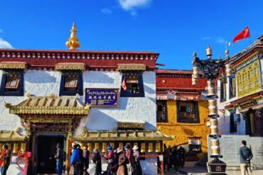 Exterior of a Buddhist monastery with white walls, ornate gold-trimmed doorway, and a row of gold prayer wheels along the front; visitors line up in the courtyard under a bright blue sky.Barkhor Street,Lhasa,Tibet,Chinatravel