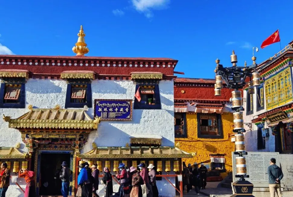 Exterior of a Buddhist monastery with white walls, ornate gold-trimmed doorway, and a row of gold prayer wheels along the front; visitors line up in the courtyard under a bright blue sky.Barkhor Street,Lhasa,Tibet,Chinatravel