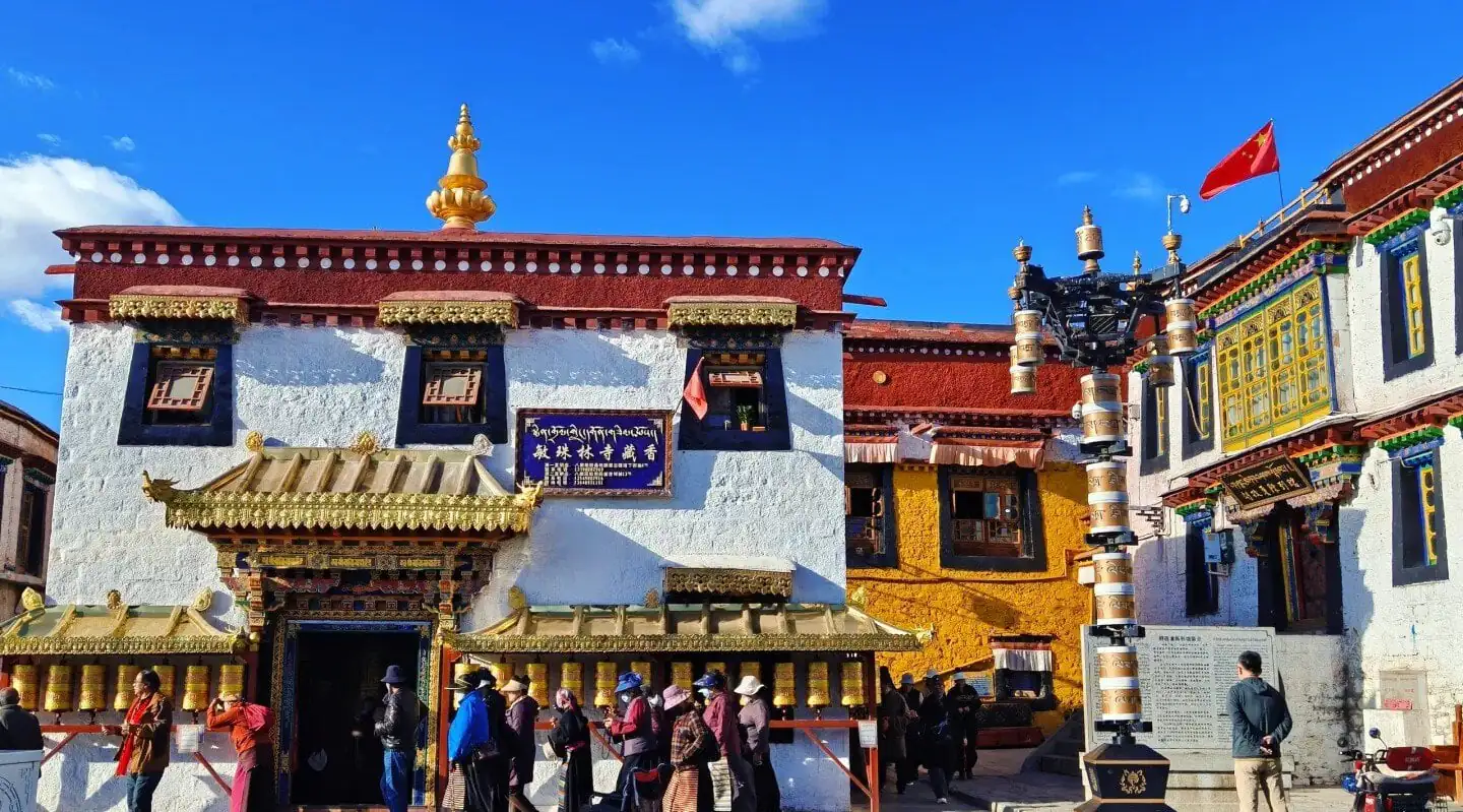 Exterior of a Buddhist monastery with white walls, ornate gold-trimmed doorway, and a row of gold prayer wheels along the front; visitors line up in the courtyard under a bright blue sky.Barkhor Street,Lhasa,Tibet,Chinatravel