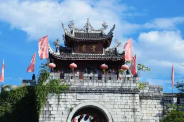 Traditional Chinese temple gate atop a stone archway with red lanterns and pink banners against a blue sky.Qingyan Ancient Town,Guiyang,Guizhou