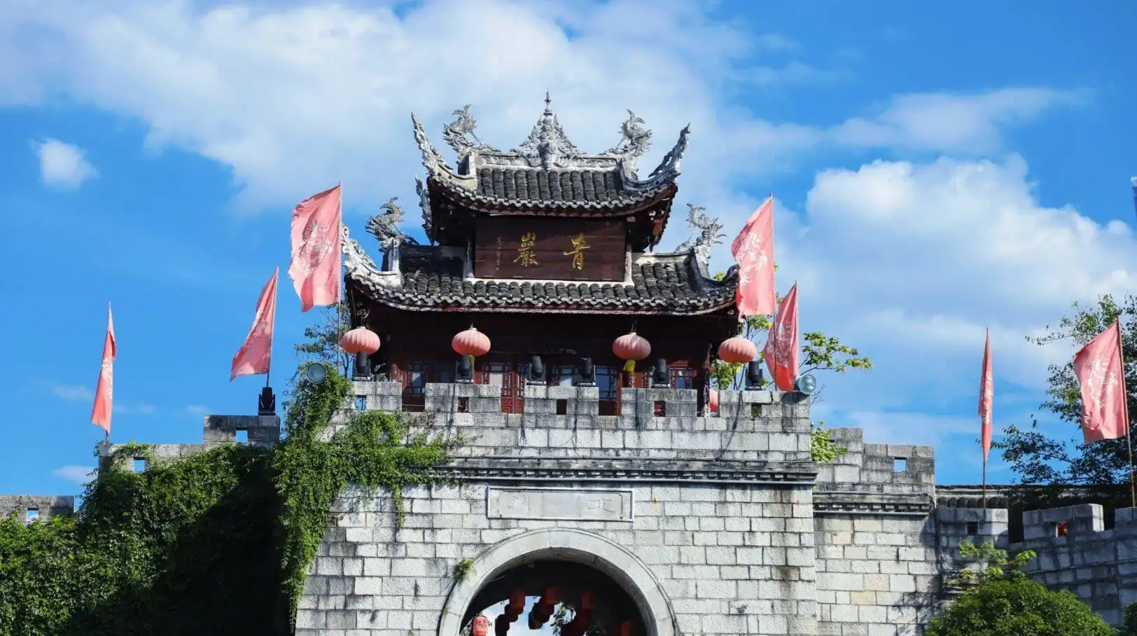 Traditional Chinese temple gate atop a stone archway with red lanterns and pink banners against a blue sky.Qingyan Ancient Town,Guiyang,Guizhou