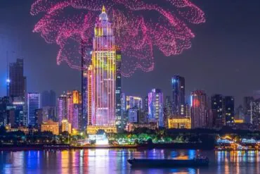Nighttime city skyline with a tall illuminated central skyscraper, pink fireworks overhead, and reflections on the river with a boat in the foreground.Yangtze River Cruise,Wuhan,Hubei ,Chinatravel