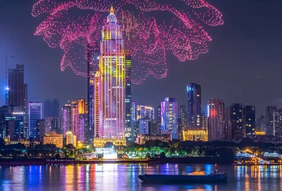 Nighttime city skyline with a tall illuminated central skyscraper, pink fireworks overhead, and reflections on the river with a boat in the foreground.Yangtze River Cruise,Wuhan,Hubei ,Chinatravel