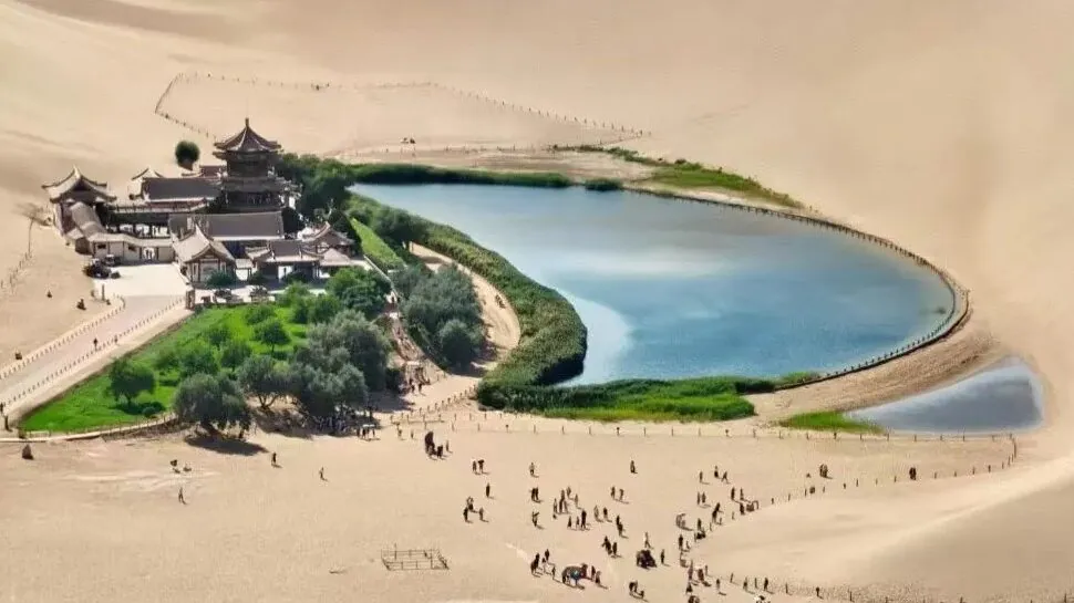 Desert oasis with a circular blue lake surrounded by green lawns and a traditional Chinese temple complex nestled in sand dunes.Crescent Lake & Mingsha Mountain,Dunhuang,Gansu