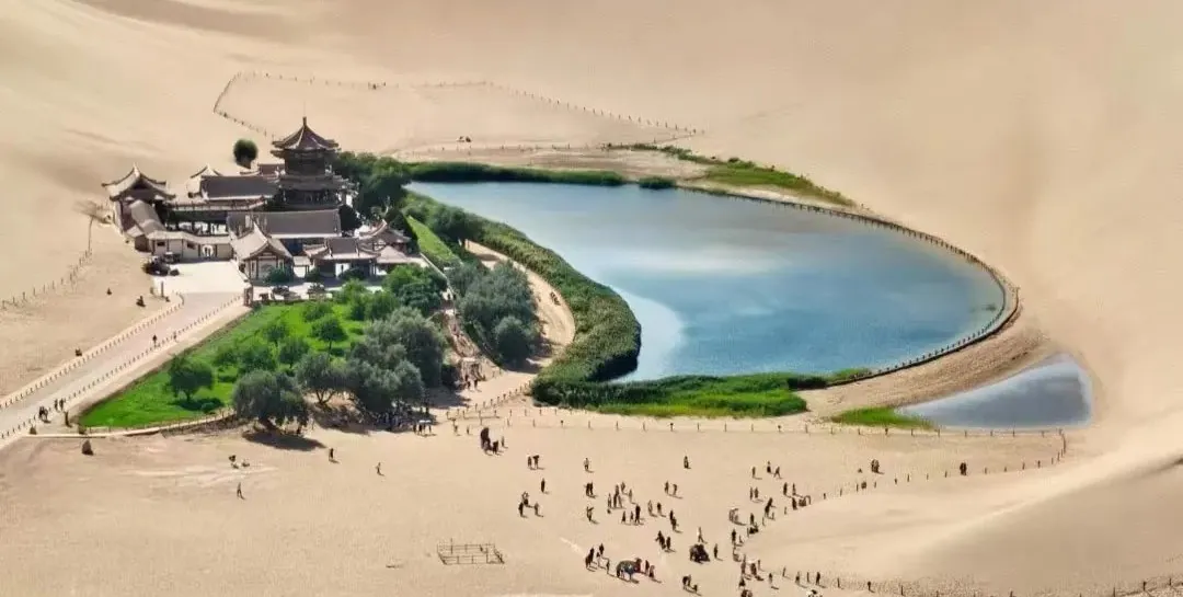 Desert oasis with a circular blue lake surrounded by green lawns and a traditional Chinese temple complex nestled in sand dunes.Crescent Lake & Mingsha Mountain,Dunhuang,Gansu