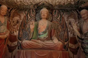 Central seated Buddha statue in a richly painted cave temple, with two standing figures on either side.Dunhuang Museum,Dunhuang,Gansu