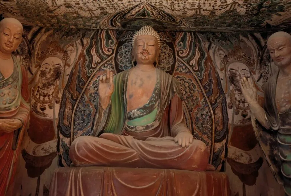 Central seated Buddha statue in a richly painted cave temple, with two standing figures on either side.Dunhuang Museum,Dunhuang,Gansu
