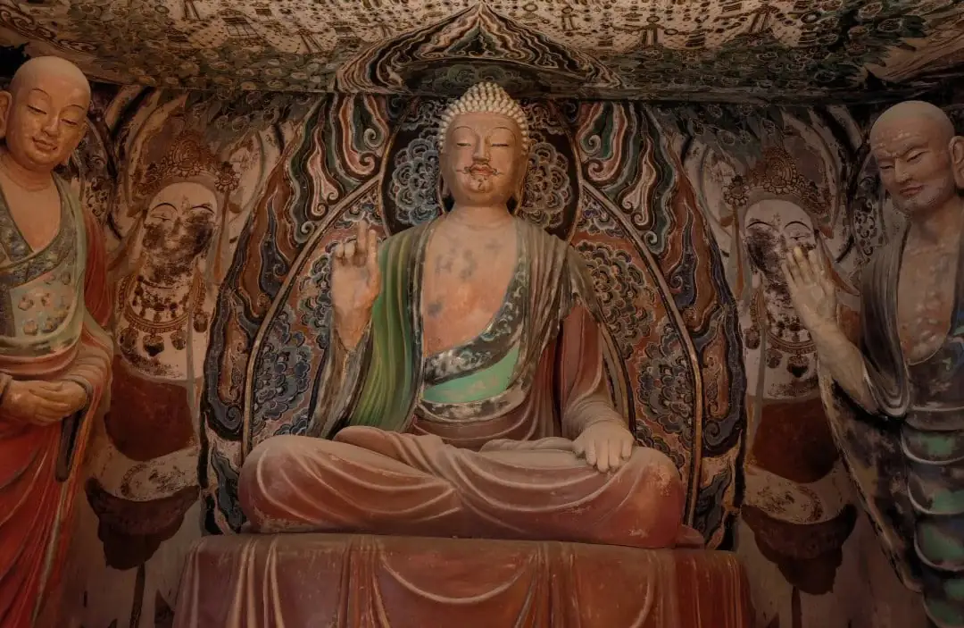 Central seated Buddha statue in a richly painted cave temple, with two standing figures on either side.Dunhuang Museum,Dunhuang,Gansu