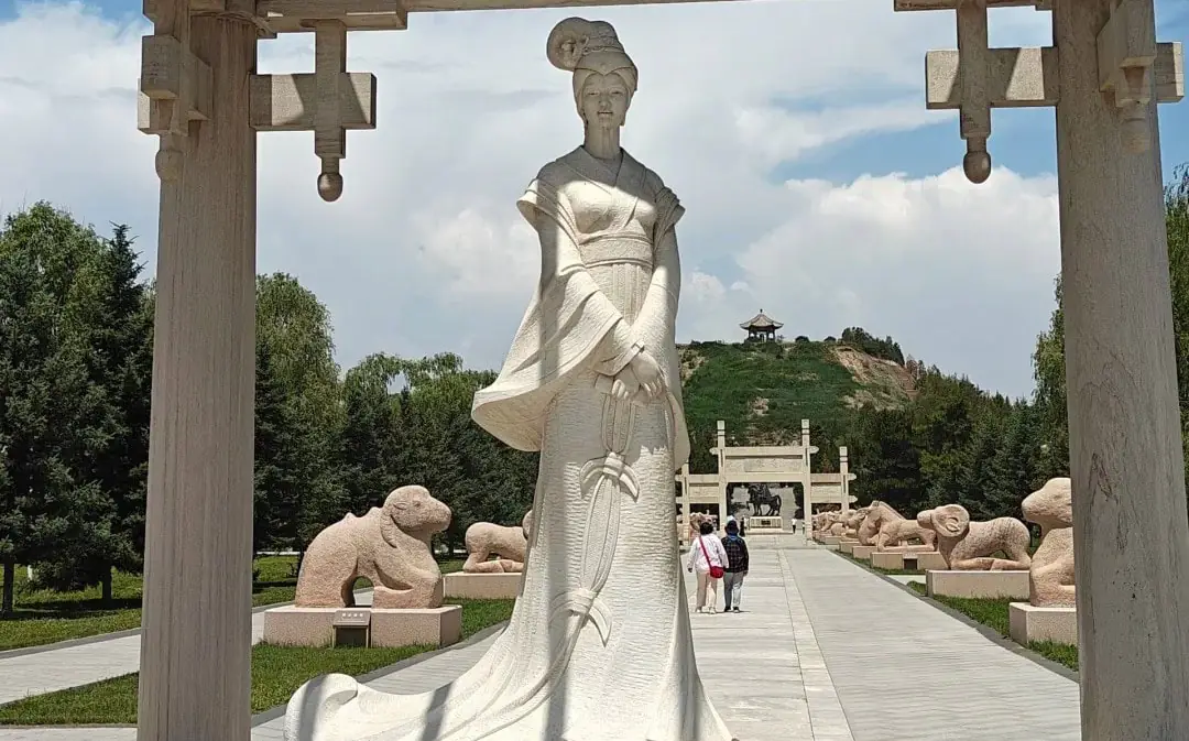 Large white statue of a woman in flowing traditional dress standing along a stone walkway, with stone animal sculptures lining the path and a hilltop pavilion in the distance.Zhaojun Tomb,Hohhot,Inner Mongolia,Chinatravel