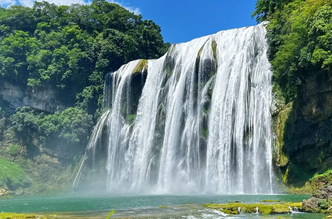 Waterfall cascades down a rocky cliff into a turquoise pool, surrounded by lush green forest on a sunny day.Huangguoshu Waterfall,Guiyang,Guizhou