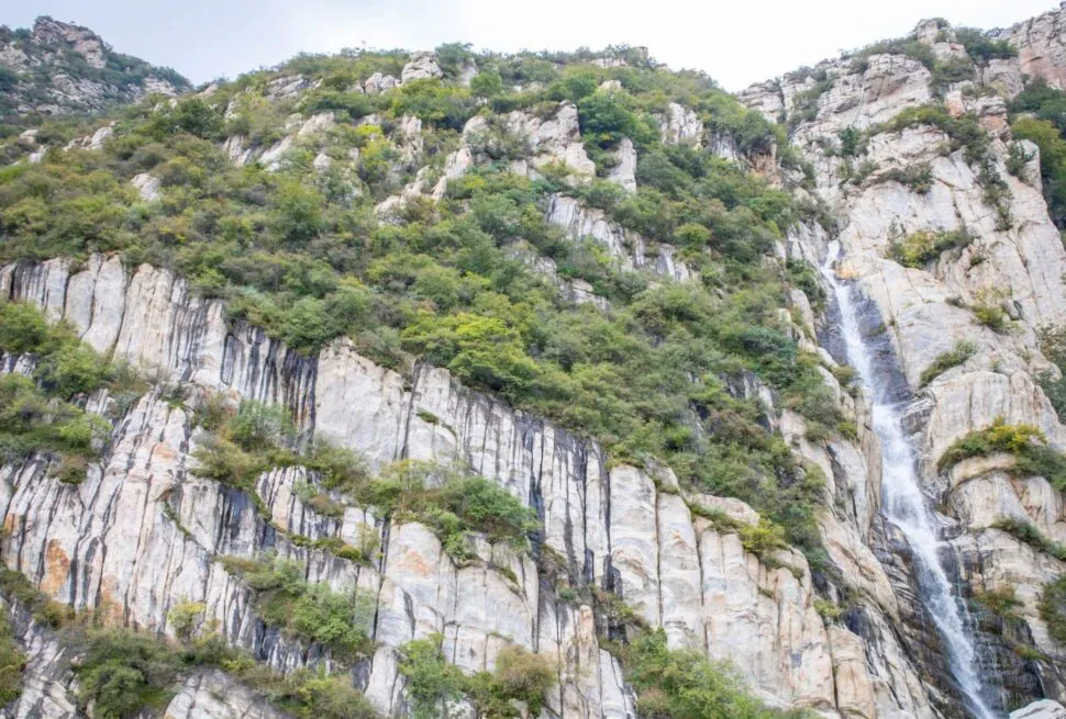 Steep rocky mountainside with striped vertical rock faces, green shrubs, and a narrow waterfall on the right side.Songshan Luya Waterfall,Shaolin & Songshan Area,Henan,Chinatravel