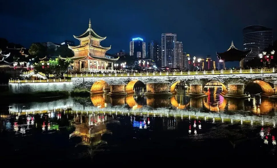 Night scene of a lit traditional Chinese pagoda and arched bridge over a calm river with reflections in the water.Jiaxiu Tower,Guiyang,Guizhou