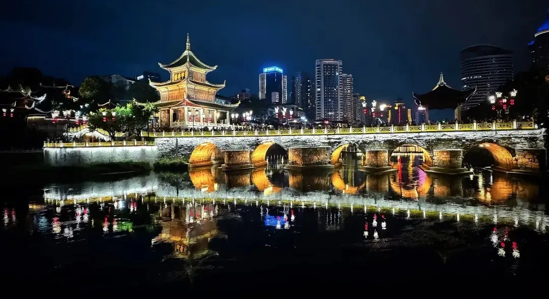 Night scene of a lit traditional Chinese pagoda and arched bridge over a calm river with reflections in the water.Jiaxiu Tower,Guiyang,Guizhou