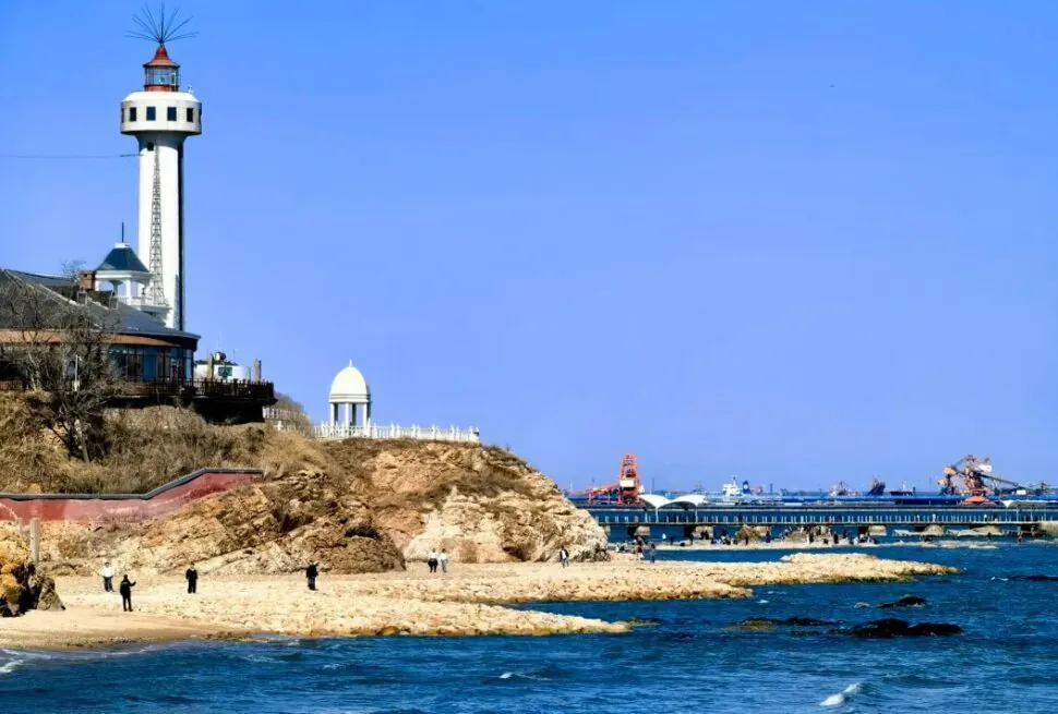 Lighthouse atop a rocky cliff overlooking a sandy beach and calm blue sea with a pier in the distance under a clear sky.Beidaihe,Qinhuangdao,Hebei,Chinatravel