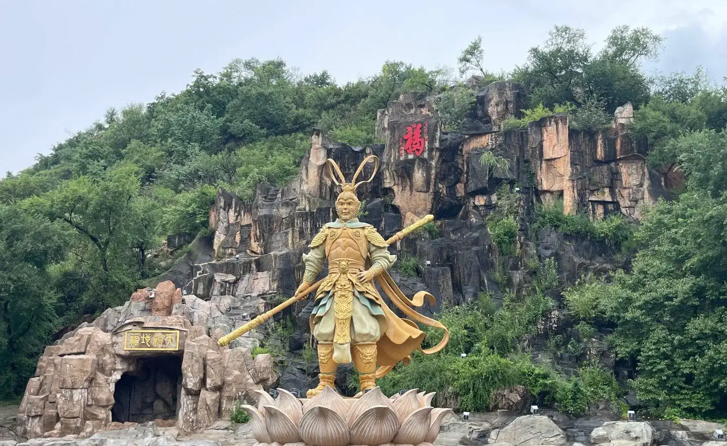Golden armored warrior statue holding a long staff, standing on a lotus pedestal at a rocky hillside with greenery and a cave entrance behind him.Jilin Beishan Park,Chinatravel