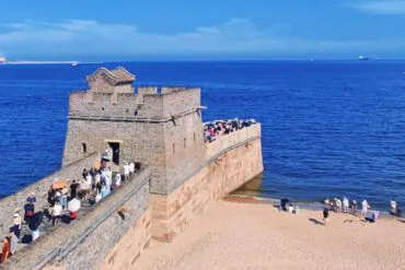 Stone coastal fortress with a walkway filled with visitors along the wall, overlooking a calm blue sea on a sunny day.Shanhaiguan,Qinhuangdao,Hebei,Chinatravel