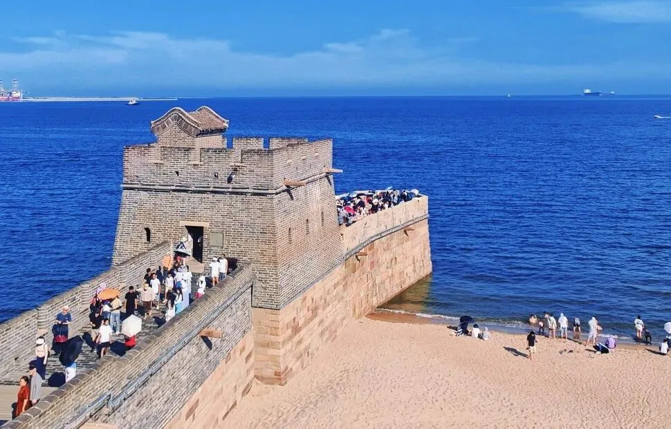Stone coastal fortress with a walkway filled with visitors along the wall, overlooking a calm blue sea on a sunny day.Shanhaiguan,Qinhuangdao,Hebei,Chinatravel