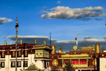 Golden Tibetan temple complex with gilded rooftops, prayer wheels, and decorated poles against a bright blue sky with scattered clouds,Jokhang Temple,Lhasa,Tibet,Chinatravel