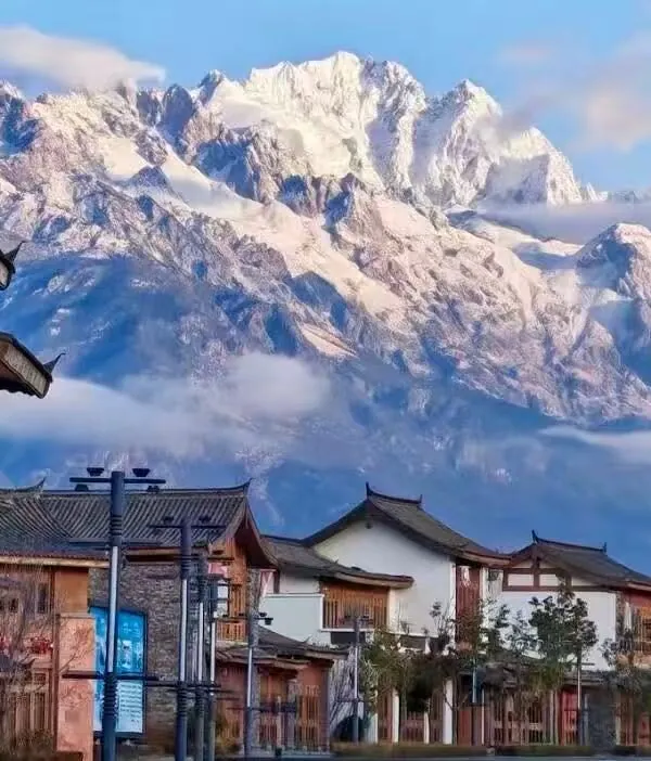 Snow-capped mountains tower over a street of traditional wooden buildings, blue sky and light clouds above.Jade Dragon Snow Mountain,Lijiang,Yunnan,Chinatravel