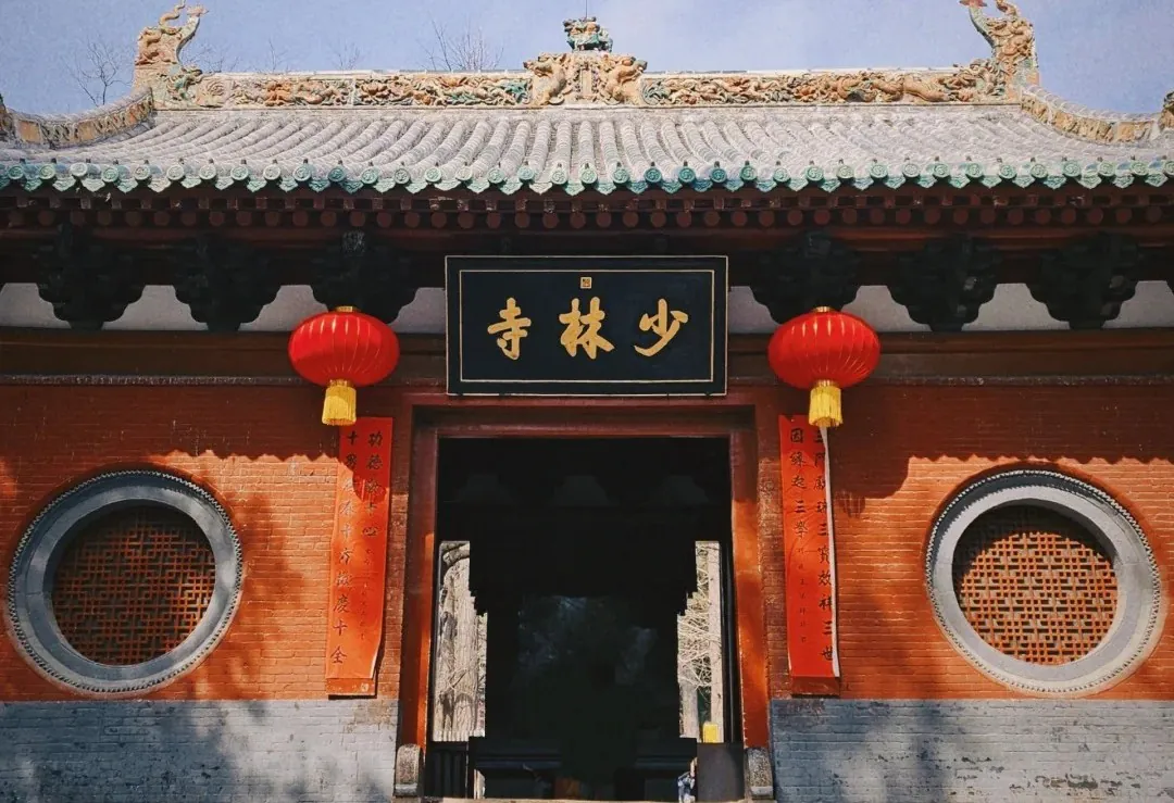 Entrance to a Shaolin Temple with red brick walls, round lattice windows, red lanterns, and a black plaque with gold Chinese characters.Shaolin Temple,Shaolin & Songshan Area,Henan,Chinatravel